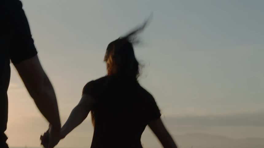 Silhouette of a young couple running while holding hands on the sand next to the ocean at sunset