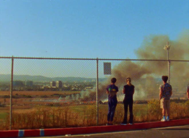 Three teenagers stand behind a fence and watch the smoke from a crashed vehicle