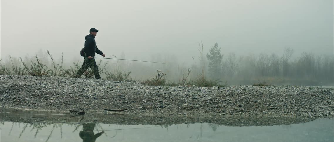 Man with black cap coat and pants walks along river bank carrying fishing rod on cloudy and foggy day