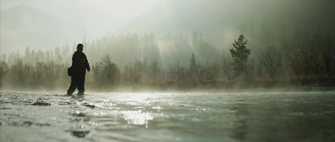 A fisherman casts his fishing line while standing in a river on a misty morning