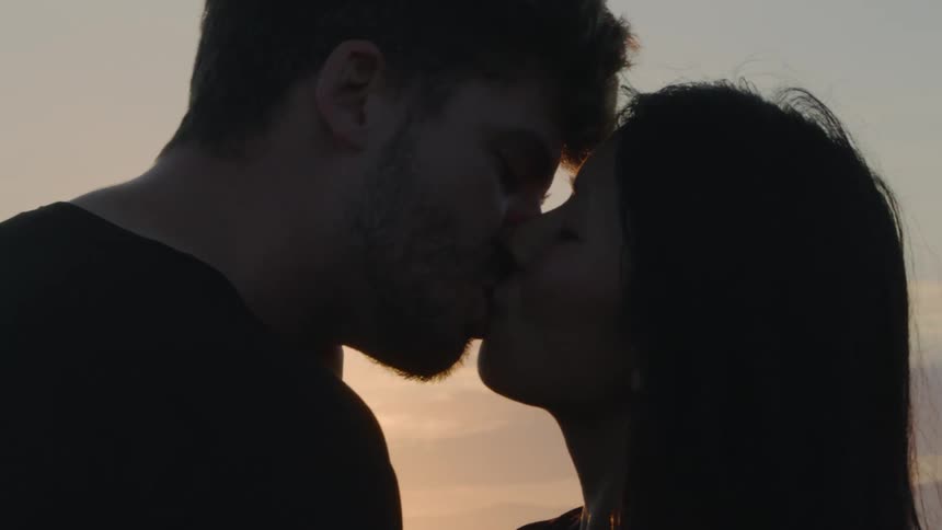 Sunlight shines behind the silhouette of a young couple kissing at sunset