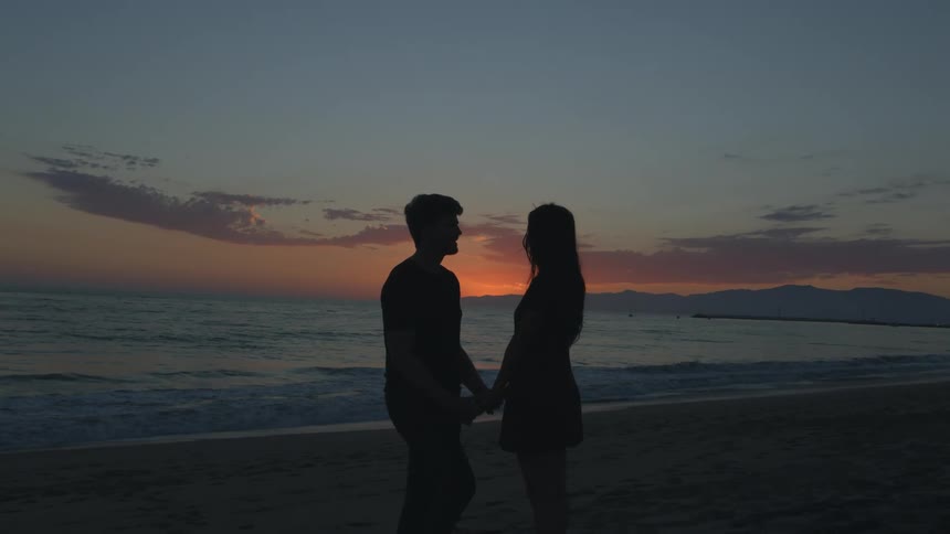 A man and woman stand on the beach and hold hands as they face one another and lean forwards and kiss