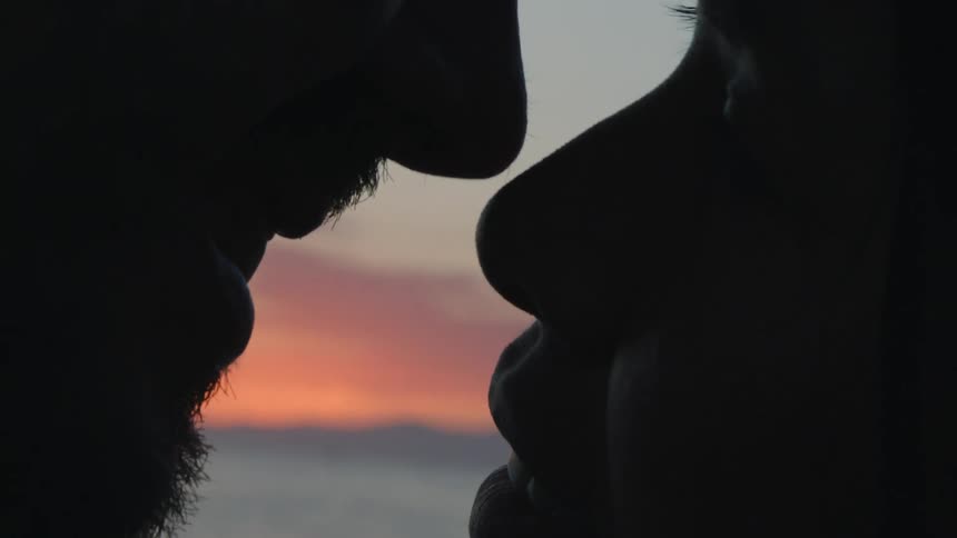 Man with beard and woman making out are silhouetted against sky on the beach at dusk