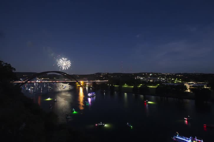 Boats are sailing in the water as traffic moves across the bridge and fireworks light up the night sky