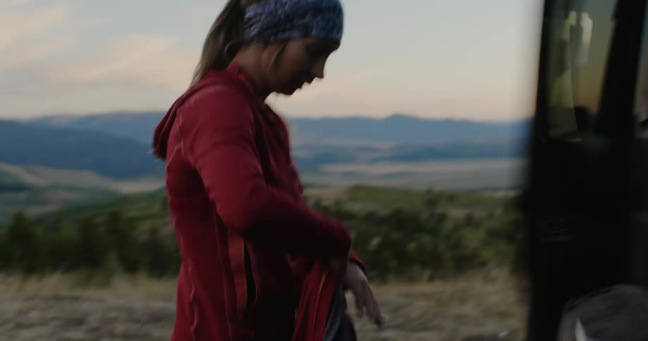 A woman retrieves a jacket from the backseat of a vehicle parked off road and walks out of frame