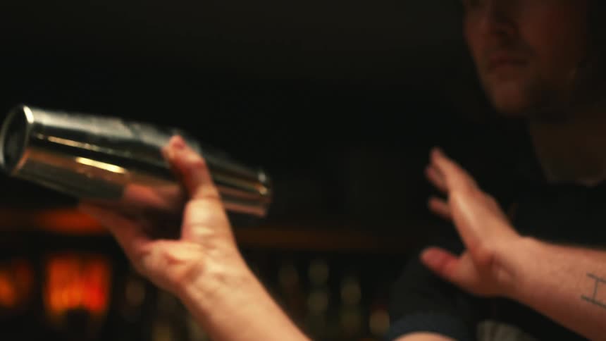 Barman with black t shirt gray apron and tattoo shakes cocktail in bar