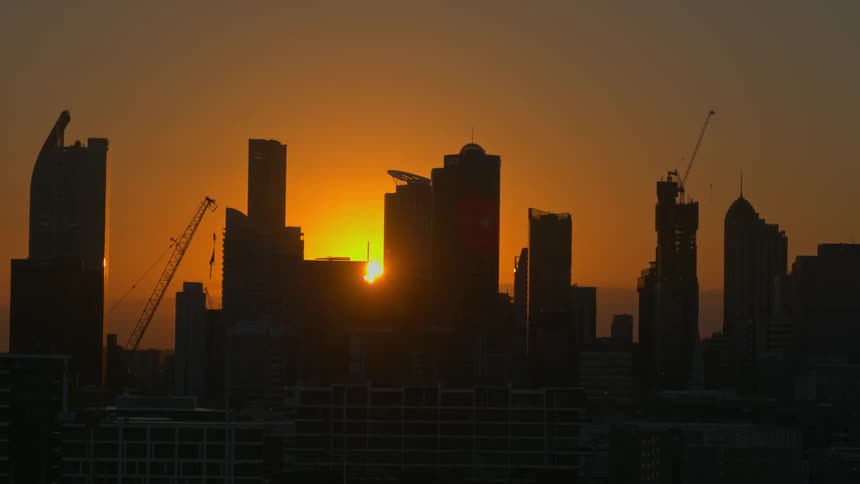 Silhouette of tall city buildings as construction cranes are working under an orange sky as the sun sets in the distance