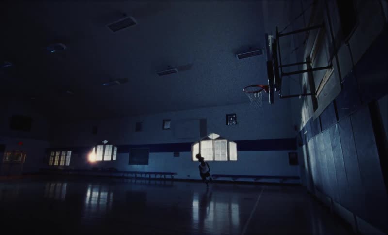 Young athletic teenager slam dunks a basketball inside a dimly lit court at morning