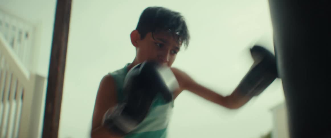 A boy wearing boxing gloves is on his porch hitting a punching bag