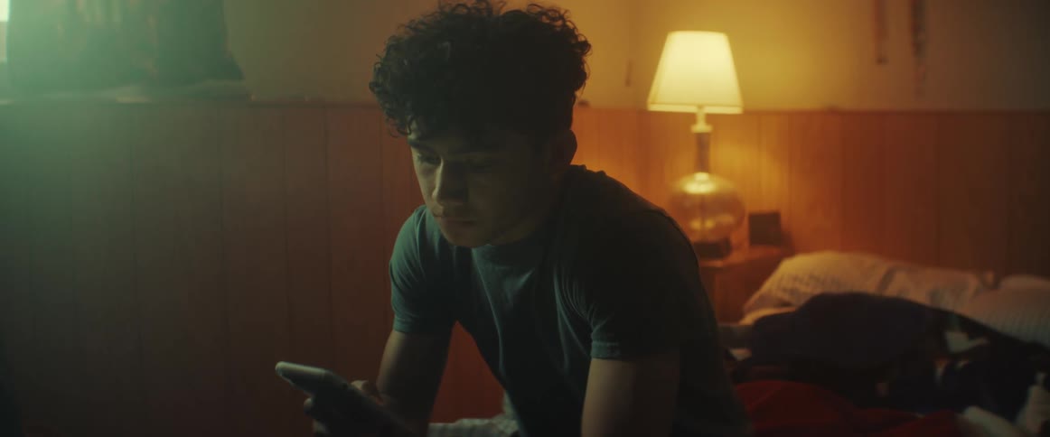 Young teenage boy checks his phone while sitting on the bed in his bedroom
