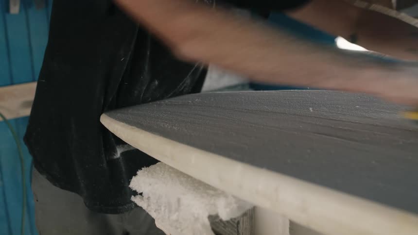 Man uses a tool to sand the surface of a surfboard inside a small workshop
