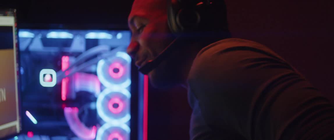 Happy relaxed man sitting in front of a computer under bright colorful rgb lights talking off a headset