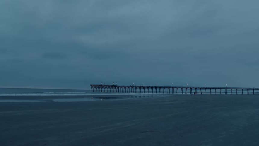 People move along beach near pier as surf rolls onto sand under an overcast sky