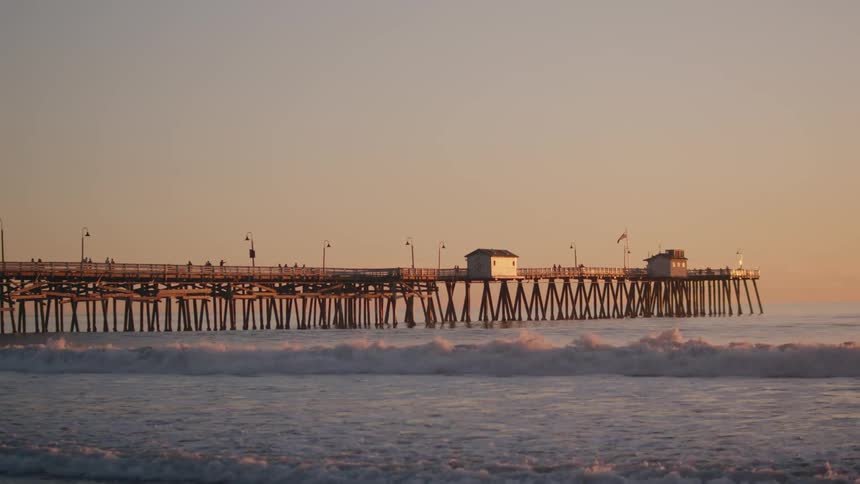 Surfer rides on waves while standing on surfboard at dusk near the long pier