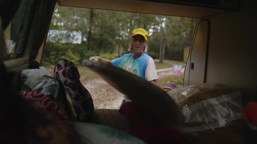 Woman with yellow cap gray hair and pink blue t shirt takes surfboard from car