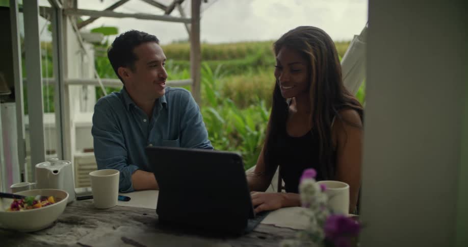A man and a woman are sitting outside on a patio talking as she types on a laptop computer