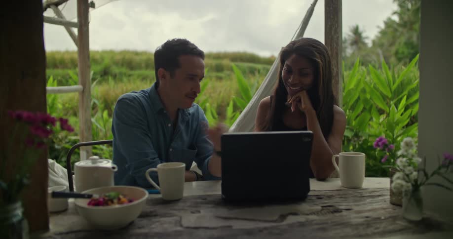 A man points at a laptop and gestures as he talks with a coworker at a meeting on a patio