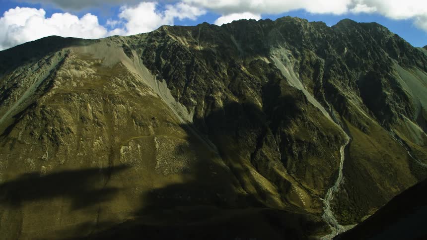 Over a grass covered mountain is a mountain ridge peaking high in a cloud filled sky