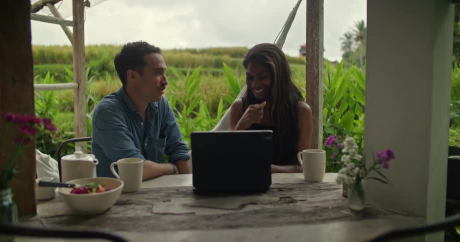 Man with mustache blue shirt and woman with brown hair and black top sit at table working on tablet smiling in the porch