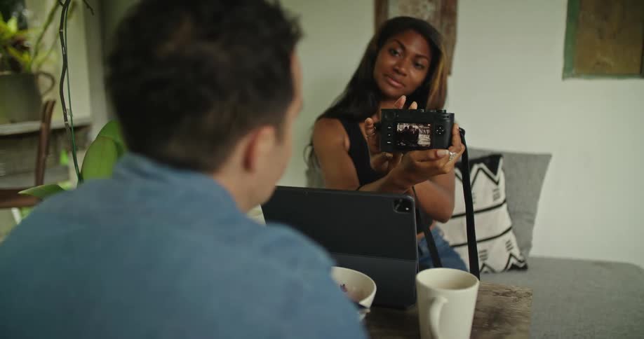 A man and a woman are sitting in a room together and she shows him a picture on her camera as she is sitting behind a laptop computer