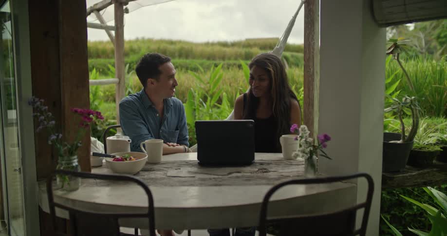 Young couple sitting on a small table in the porch of a tropical house working on a laptop