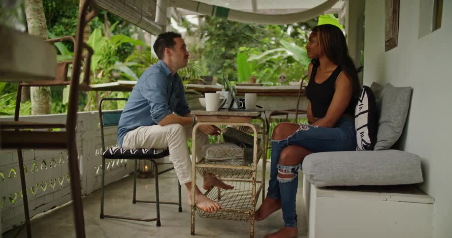 Man with mustache blue shirt and beige pants sits at table talking to woman with brown hair black top and ripped jeans on the balcony
