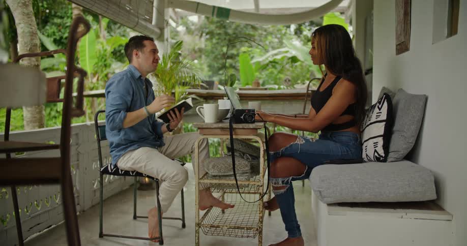 Young couple sitting on a small table in the porch of a tropical house working on laptops and reading books