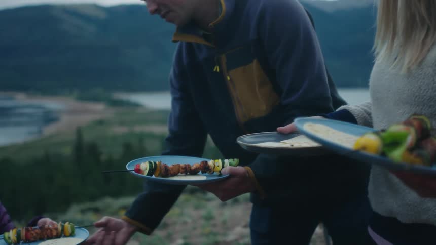 A man and woman carry plates of food over to their friends while camping