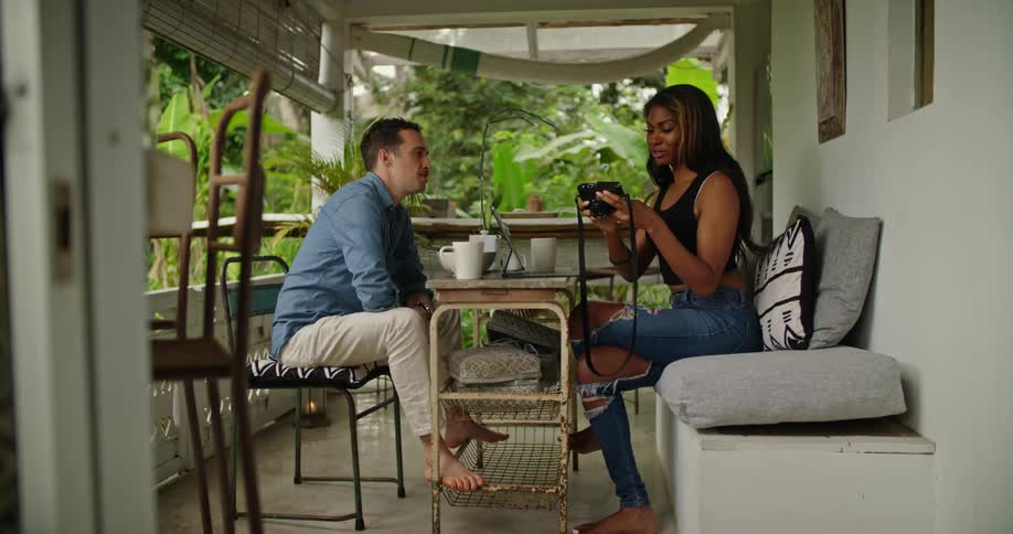 Young couple eating breakfast look at pictures on a camera sitting on the porch of a house on the tropics
