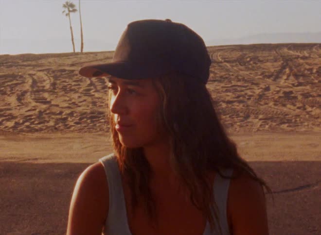 Woman with black cap wavy hair and gray top looks at camera in the desert