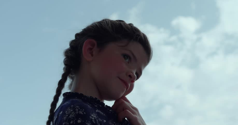 A little girl with her long hair in braids smiles and turns around as she stands under a cloudy sky