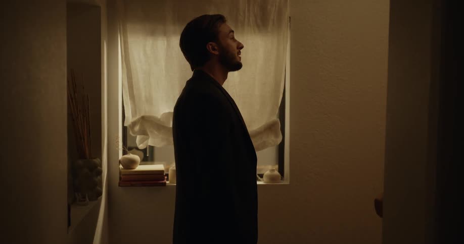 Man following a woman inside a house stops for a moment at the stairs next to a window