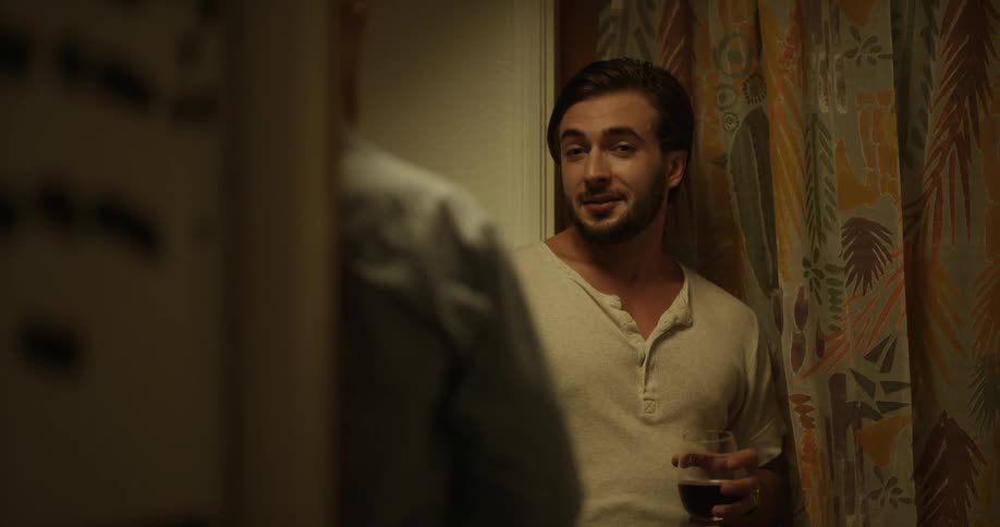 Man with beard and white t shirt holds a glass of wine talks to person smiling