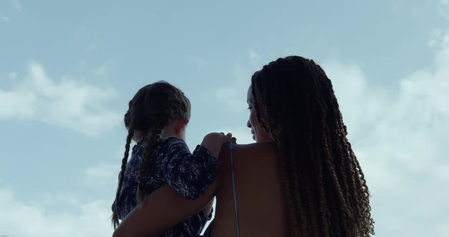 A woman is holding her little girl and they look at each other and smile under a cloudy sky