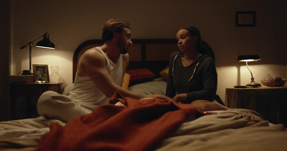Man with beard white undershirt and gray pants talks with woman with dreadlocks black shirt and shorts on bed in bedroom