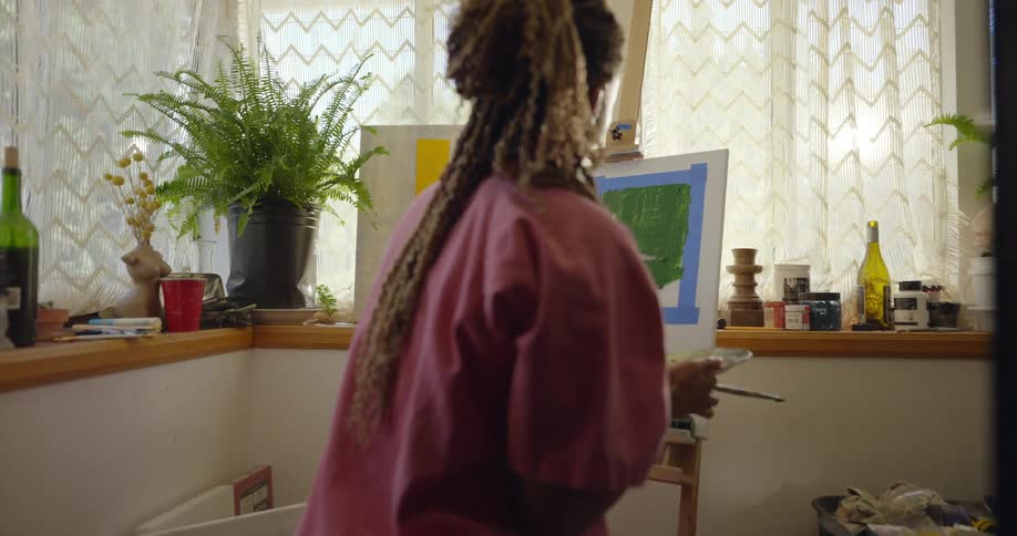 A woman is sitting in a room with a lot of windows as she paints on a canvas