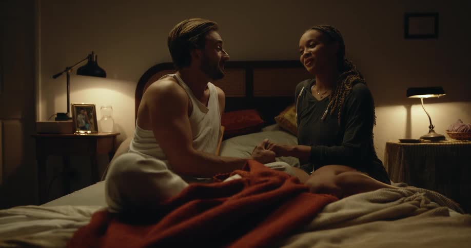 Man with beard white undershirt and gray pants holding hands and talking makes out with woman with dreadlocks black shirt and shorts on bed in bedroom