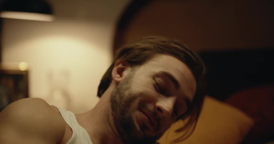Man with beard white undershirt and gray pants talks and makes out with woman with dreadlocks black shirt and shorts on bed in bedroom