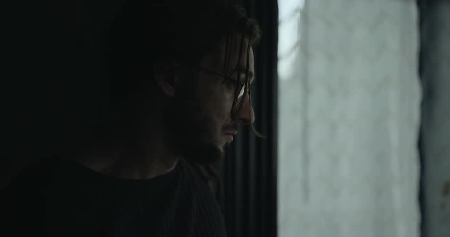 Young bearded man with long hair wearing glasses looks pensively out the window