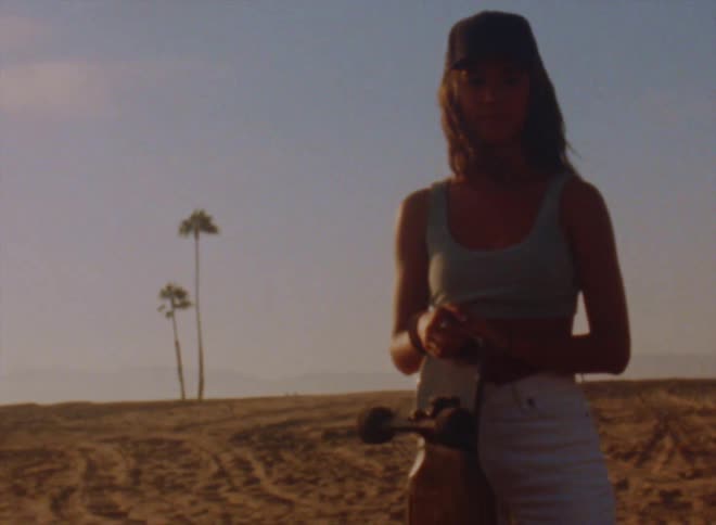 Woman with black cap wavy hair blue top and white pants holds skateboard posing for camera in barren desert