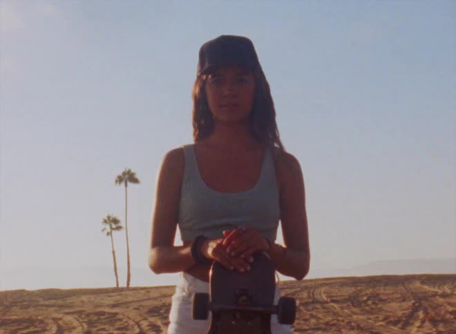 A teenage girl wearing a cap is standing on a beach with her skateboard