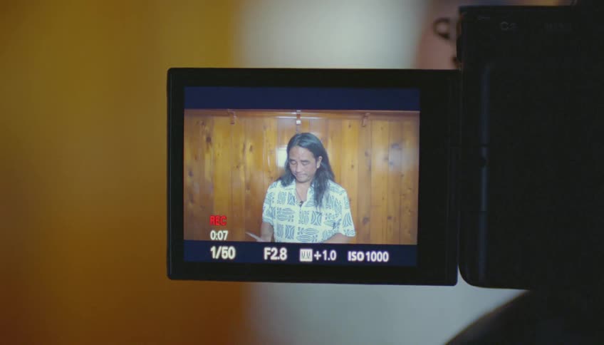 Man with long hair and white shirt films himself with camera reading a piece of paper