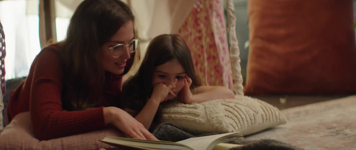 Mother with glasses brown hair and red sweater reads book with daughter lying next to her in living room at home