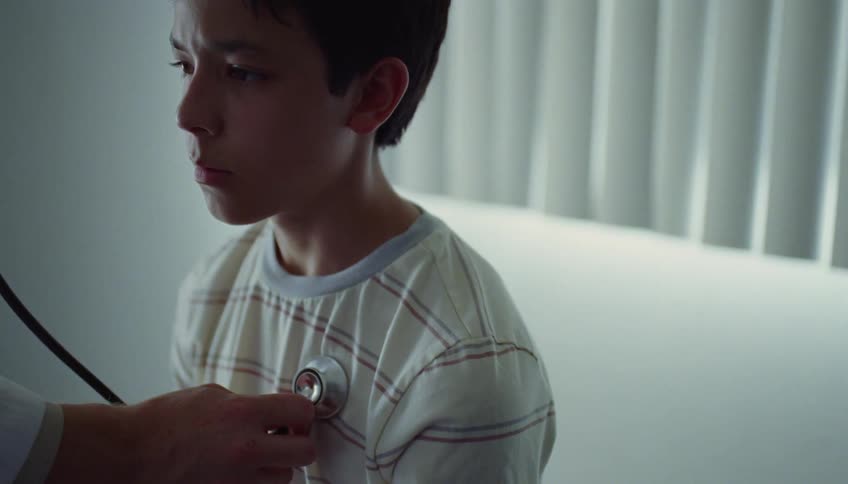 A boy is sitting in a doctors office with a worried look on his face as the doctor puts his stethoscope to the boys chest