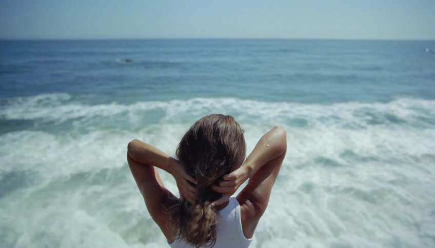 A woman is standing along the coast running her hands through her long hair as she watches the waves roll in to shore