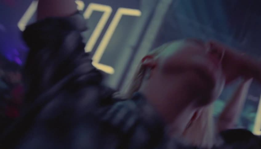 A group of men and women dance at a club as lights flash and their friend carries drinks to them