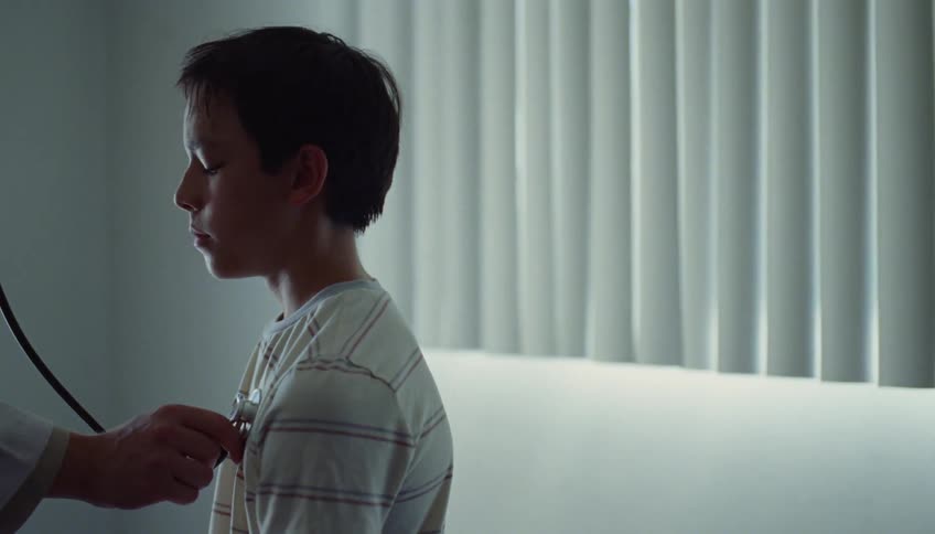 A doctor holds a stethoscope to a boys chest in an exam room