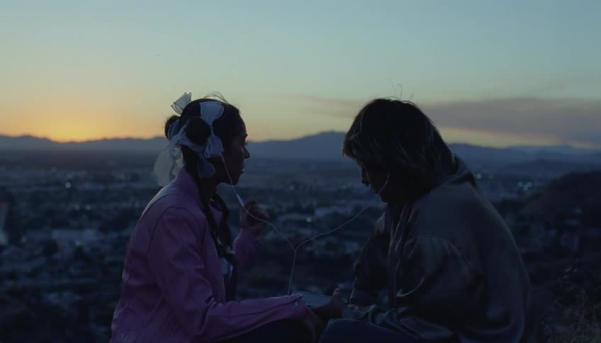 A man and a woman are sitting outside and hes holding a phone and they each have an earplug in their ear with the city below and mountains in the distance