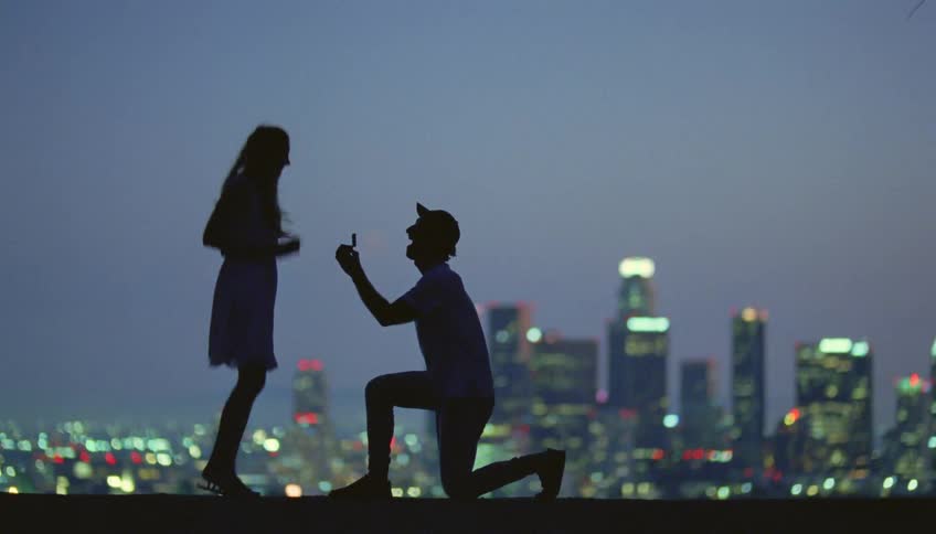 The silhouette of a man getting down on one knee proposing and a woman jumping for joy