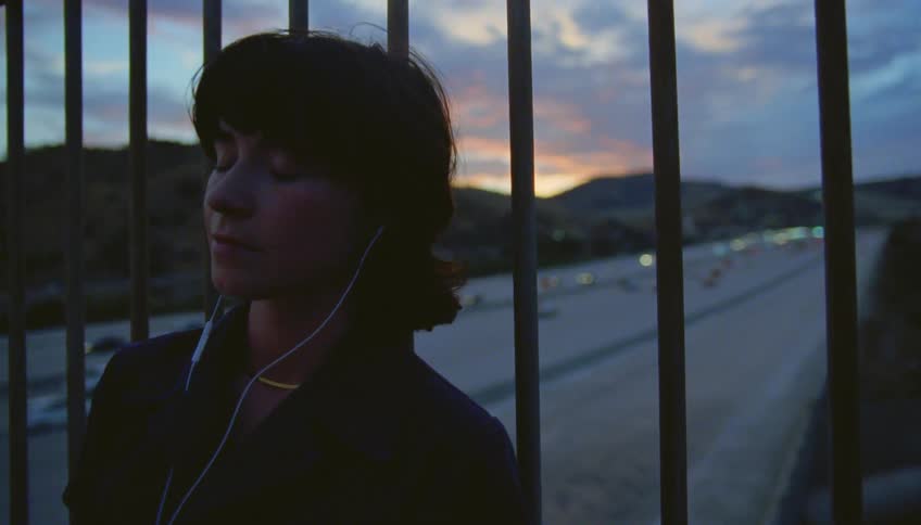 A teenage girl is standing on a bridge leaning against the fence and listening to music in her headphones as traffic moves on the freeway below